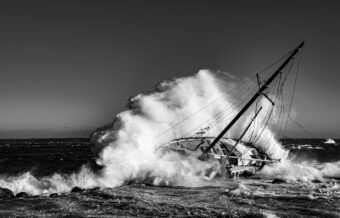 Voilier pris dans une mer démontée, vagues se brisant sur la coque, en noir et blanc